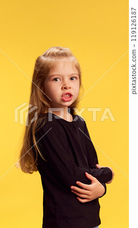Little rude girl with long blonde hair crosses her arms while pouting in frustration against yellow studio background. 119126187