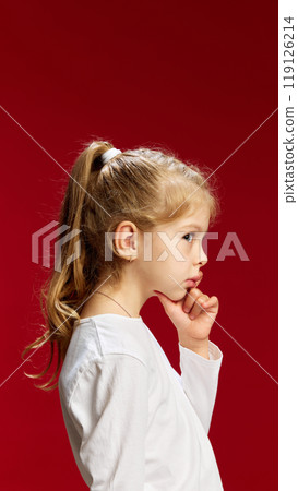Looking away from camera, little girl smiles with gentle expression, highlighting her playful and joyful mood against red studio background. Looking away from camera, little girl smiles with gentle expression, highlighting her playful and joyful mood against red studio background. 119126214