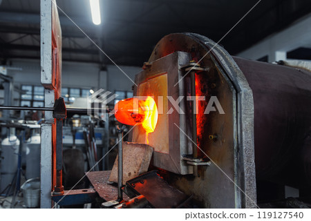 Close-up glassblowing process tube tool with molten glass shaped inside refractory furnace in industrial craft workshop. Handmade blowing glassware design production manufacture Europe Close-up glassblowing process tube tool with molten glass shaped inside refractory furnace in industrial craft workshop. Handmade blowing glassware design production manufacture Europe 119127540