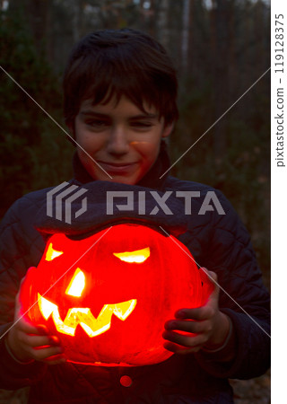 A joyful kid with a big smile holds a Halloween pumpkin while surrounded by a spooky, atmospheric forest setting 119128375