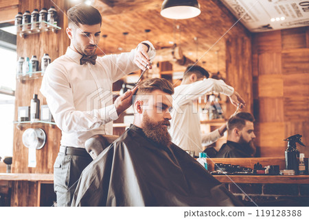 Everything should be perfect. Side view of young bearded man getting haircut by hairdresser while sitting in chair at barbershop Everything should be perfect. Side view of young bearded man getting haircut by hairdresser while sitting in chair at barbershop 119128388