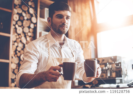 Here you go! Young handsome man in apron holding two coffee cups and looking away while standing at cafe Here you go! Young handsome man in apron holding two coffee cups and looking away while standing at cafe 119128756