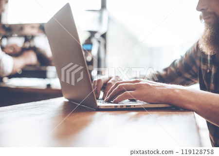 Surfing net at cafe. Close-up part of young bearded man using his laptop while sitting at bar counter at cafe with barista at the background Surfing net at cafe. Close-up part of young bearded man using his laptop while sitting at bar counter at cafe with barista at the background 119128758