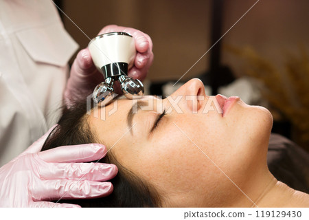 Cosmetologist performing phonophoresis ultrasound therapy on woman's face 119129430