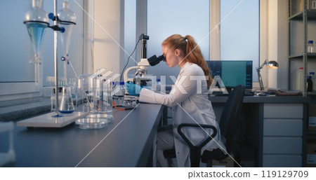 Young female scientist sits in chair, looks under microscope and examines samples Young female scientist sits in chair, looks under microscope and examines samples 119129709