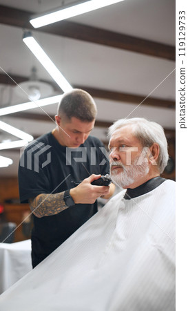 Barber using electric clippers to clean up sides of the man hair, focusing on neatness and symmetry. Senior man visiting barbershop. 119129733