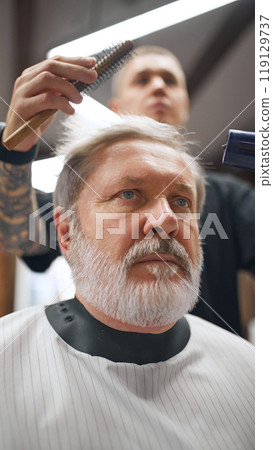 Barber shaping man hair with round brush and hairdryer, emphasizing detail and technique. Elderly man making hairstyle in barber shop Barber shaping man hair with round brush and hairdryer, emphasizing detail and technique. Elderly man making hairstyle in barber shop 119129737