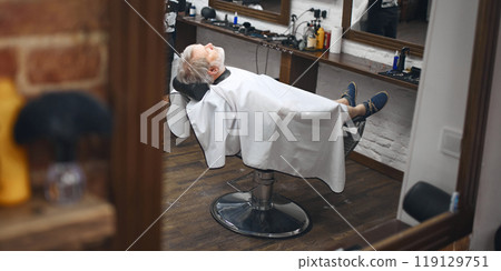 Client reclining in chair, fully relaxed, showcasing the comfort. Elderly man visiting barbershop for hairstyle 119129751