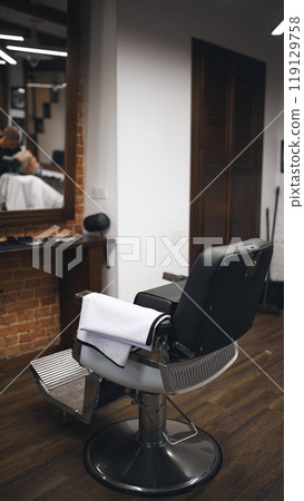 Empty barber chair with clean towel draped over armrest, awaiting client. Wooden floors and cozy barbershop atmosphere 119129758