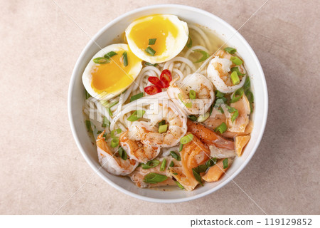 Asian dish with broth, noodles, tiger shrimp, fish, egg and green onions. Traditional Asian soup in a white deep plate. Selective focus, close-up. Asian dish with broth, noodles, tiger shrimp, fish, egg and green onions. Traditional Asian soup in a white deep plate. Selective focus, close-up. 119129852