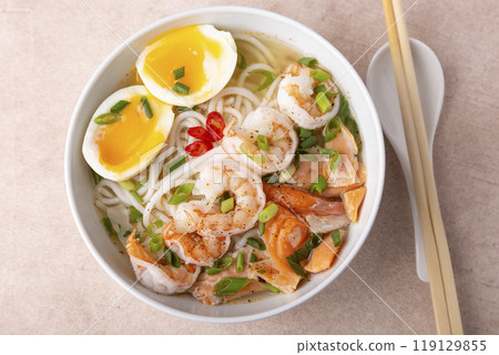 Asian dish with broth, noodles, tiger shrimp, fish, egg and green onions. Traditional Asian soup in a white deep plate. Selective focus, close-up. Asian dish with broth, noodles, tiger shrimp, fish, egg and green onions. Traditional Asian soup in a white deep plate. Selective focus, close-up. 119129855