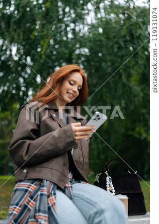 Vertical low-angle portrait of cheerful young woman with long red hair using smartphone while sitting on bench outdoors in park, enjoying fresh air and greenery, smiling looking to phone screen. Vertical low-angle portrait of cheerful young woman with long red hair using smartphone while sitting on bench outdoors in park, enjoying fresh air and greenery, smiling looking to phone screen. 119131074