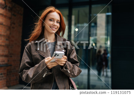 Portrait of cheerful young woman with long red hair wearing brown leather jacket standing on urban street on summer day, holding using smartphone, smiling looking at camera, enjoying free time in city 119131075