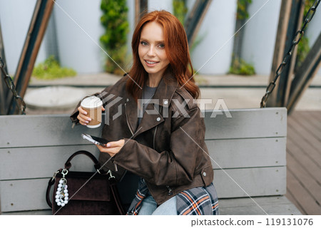Portrait of charming redhead female smiling looking at camera swinging on swings in park, drinking coffee and browsing smartphone. Happy woman enjoying coffee on swing, using typing phone Portrait of charming redhead female smiling looking at camera swinging on swings in park, drinking coffee and browsing smartphone. Happy woman enjoying coffee on swing, using typing phone 119131076