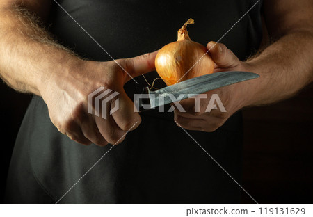 A cook cuts an onion to add to food. Close-up of the chef hands holding an onion and a knife 119131629