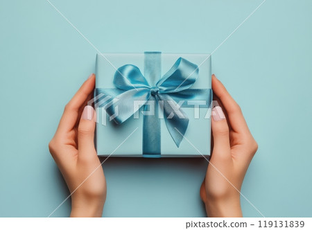 Hands holding a beautifully wrapped blue gift box with a satin ribbon on a light blue background 119131839