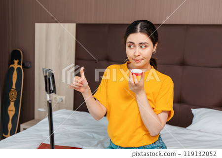 Portrait of pretty female vlogger streaming a live video to her smartphone sitting on bed and thumbs up showing cosmetics. Vlogging 119132052