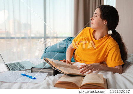 Portrait of young caucasian woman is lying on her bed and holding a book. Laptop and documents are lying next to each other. The concept of online courses and online education Portrait of young caucasian woman is lying on her bed and holding a book. Laptop and documents are lying next to each other. The concept of online courses and online education 119132057
