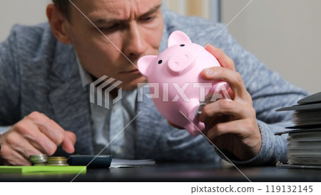 Man leans over his desk, inspecting a pink piggy bank as he manages his personal finances, surrounded by papers and a calculator Man leans over his desk, inspecting a pink piggy bank as he manages his personal finances, surrounded by papers and a calculator 119132145