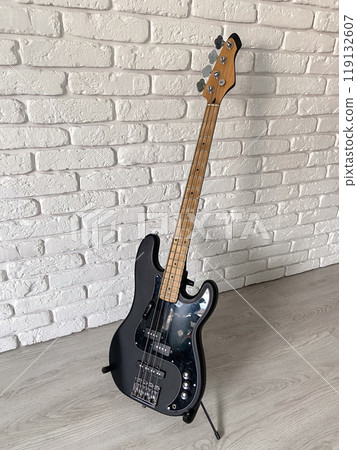 Sleek black electric bass guitar stands poised against a textured white brick wall, capturing the essence of modern music and style. The contrast between the dark instrument and the light surroundings 119132607