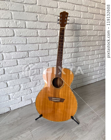 Wooden acoustic guitar stands elegantly against a textured white brick wall, creating a harmonious blend of music and art. Instrument craftsmanship 119132608