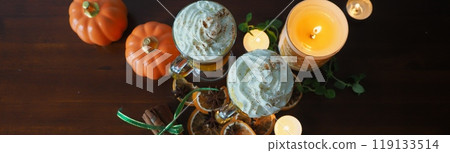 Two spicy pumpkin lattes with a cloud of whipped cream and chocolate in a cozy setting among candles, cinnamon and star anise on a wooden brown background. The mood of a warm autumn evening, the drawi 119133514