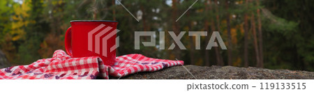Banner Red cup with a hot drink and steam on a stone and a cozy closet towel with forest motifs against a background of autumn trees of pines, birches 119133515