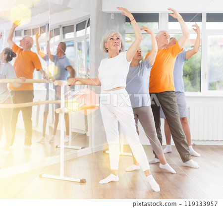 Active mature woman practicing battement tendu holding by ballet barre together with others 119133957