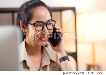 Young asian business woman using desktop computer while talking with smart phone in home office. Young asian business woman using desktop computer while talking with smart phone in home office. 119139593