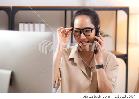 Young asian business woman using desktop computer while talking with smart phone in home office. Young asian business woman using desktop computer while talking with smart phone in home office. 119139594