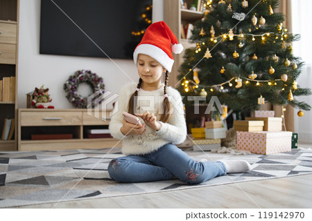 Little girl in Santa hat using phone by Christmas tree Little girl in Santa hat using phone by Christmas tree 119142970
