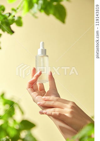 A hand delicately holds a clear glass bottle with a dropper mockup on the backdrop, which is a soft pale yellow with vibrant green leaves framing the bottle, showcasing a natural skincare product. A hand delicately holds a clear glass bottle with a dropper mockup on the backdrop, which is a soft pale yellow with vibrant green leaves framing the bottle, showcasing a natural skincare product. 119143819