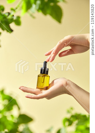 A mockup of a yellow glass bottle with a dropper that holds an essential is carefully held by a model hand in the surrounding greenery and the whitish background of the picture. A mockup of a yellow glass bottle with a dropper that holds an essential is carefully held by a model hand in the surrounding greenery and the whitish background of the picture. 119143820