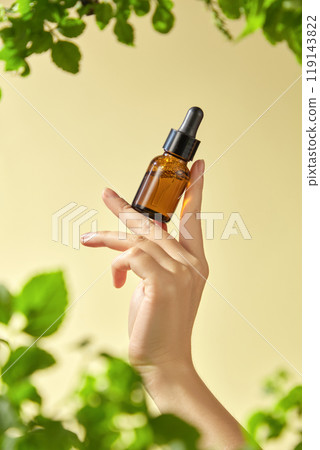 The image features a hand delicately holding a brown glass bottle with a dropper mockup, containing an essential inside. The yellow background and surrounding green foliage. 119143822
