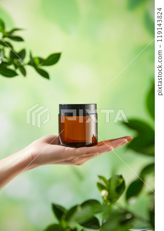 A brown glass jar with a black lid positioned in the center of the hand of the model on the outdoor background is a blurry green foliage, perfect for template advertisement of beauty care products. A brown glass jar with a black lid positioned in the center of the hand of the model on the outdoor background is a blurry green foliage, perfect for template advertisement of beauty care products. 119143824