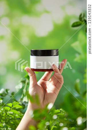 A brown glass jar with a black lid positioned in the center of the hand of the model on the outdoor background is a blurry green foliage, perfect for template advertisement of beauty care products. 119143832