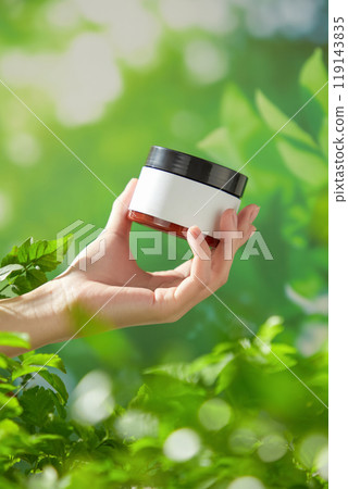 A sample photo for a beauty products ad shows a woman holding a small, brown glass jar with a black lid positioned in the center of the frame. The outdoor background is a blurry green foliage. A sample photo for a beauty products ad shows a woman holding a small, brown glass jar with a black lid positioned in the center of the frame. The outdoor background is a blurry green foliage. 119143835