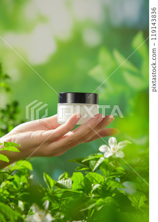 A blurred green foliage background features a model holding a small glass jar with the black cap in the center of the image, perfect for promoting natural and high-quality skincare products. 119143836