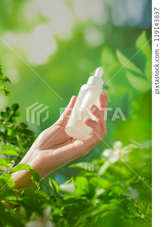 The image showcasing beauty care products features a woman hand holding an unlabeled dropper serum bottle with a white cap reaching out from the leaves in the fresh background with green bushes. 119143837