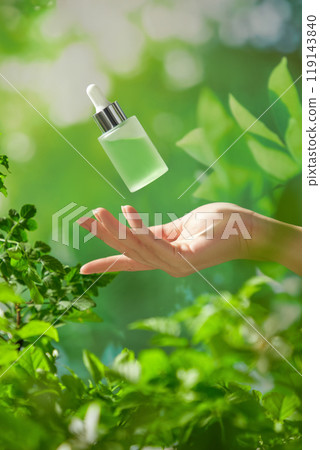 The model hand reaches out to catch an unlabeled serum bottle floating in mid-air in a lush forest filled with foliage and bright sunlight. Template image for a natural beauty product advertisement. 119143840