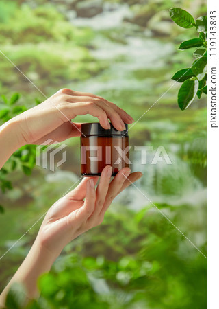 Ideal for an advertising for a beauty care product, the scenario has a stream flowing from above and fresh tree branches, and a model hand holding a brown jar mockup without a label. Ideal for an advertising for a beauty care product, the scenario has a stream flowing from above and fresh tree branches, and a model hand holding a brown jar mockup without a label. 119143843