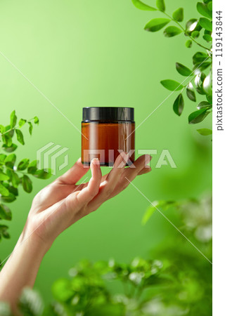 Perfect for a template commercial of beauty care goods, a hazy green foliage surrounds a brown glass jar with a black lid that is positioned in the center of the model's hand on an outdoor background. Perfect for a template commercial of beauty care goods, a hazy green foliage surrounds a brown glass jar with a black lid that is positioned in the center of the model's hand on an outdoor background. 119143844
