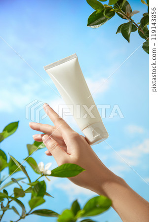 A mockup of white tube floating and touching on the back of the model arm in the blue sky background with some fresh branches. Template image for beauty care advertisement. A mockup of white tube floating and touching on the back of the model arm in the blue sky background with some fresh branches. Template image for beauty care advertisement. 119143856