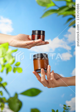 A mockup of a short and long brown jar being carried by two model hands against a background of blue skies and lush foliage is shown in a stock photo appropriate for beauty goods commercials. A mockup of a short and long brown jar being carried by two model hands against a background of blue skies and lush foliage is shown in a stock photo appropriate for beauty goods commercials. 119143866