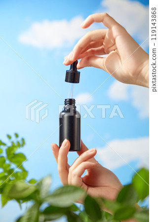 A model hand holds a glass serum bottle mockup, opening it in a blue sky background with fresh branches, promoting a natural beauty product. 119143868