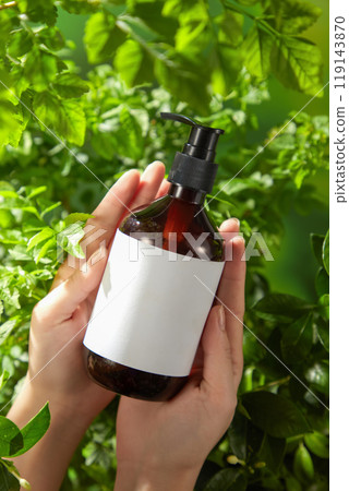 The image showcasing beauty care products features an unlabeled pump bottle lying down in the hands of the model on the leaves in the fresh background with green bushes. 119143870