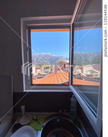 View of distant mountains framed perfectly by an open window, revealing rooftops and a clear blue sky. Indoor sink area contrasts with the vibrant outdoor scenery. 119144586