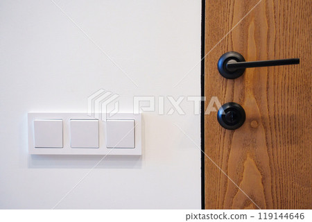 A section of a wall featuring three white light switches next to a wooden door  119144646