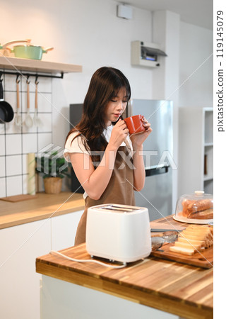 Pretty young woman drinking coffee while preparing the breakfast in the kitchen Pretty young woman drinking coffee while preparing the breakfast in the kitchen 119145079