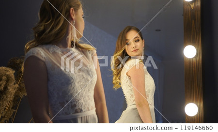Beautiful bride with long hair is wearing a white lace wedding dress and looking in a mirror Beautiful bride with long hair is wearing a white lace wedding dress and looking in a mirror 119146660